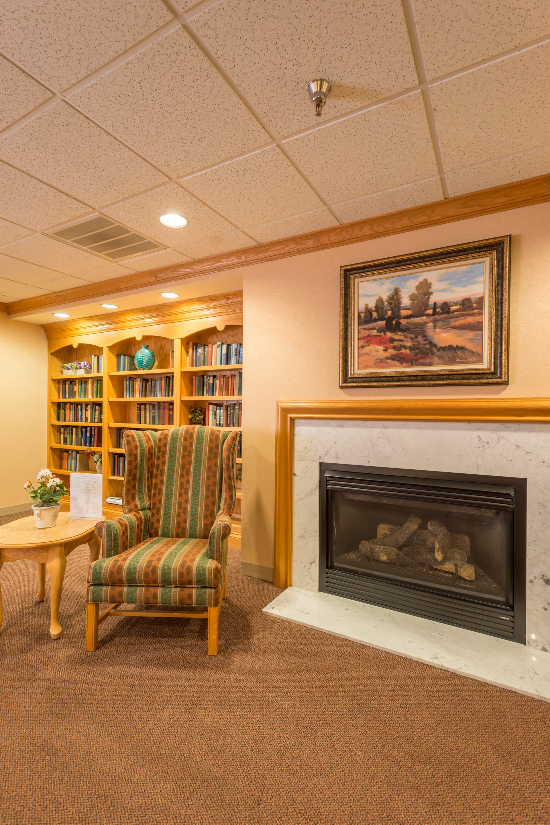 Cozy interior corner of a room featuring a striped upholstered armchair next to a wooden side table with a small potted plant and a menu or brochure. Behind the chair is a built-in wooden bookshelf filled with books and decorative items. To the right, there is a fireplace with a marble surround and a framed landscape painting hanging above it. The ceiling has recessed lighting and a sprinkler head.