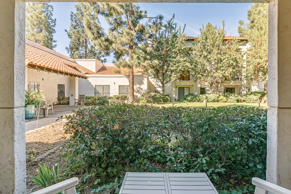 View across a landscaped courtyard with shrubs, trees, and patios toward the exterior of a multi-story senior living building.