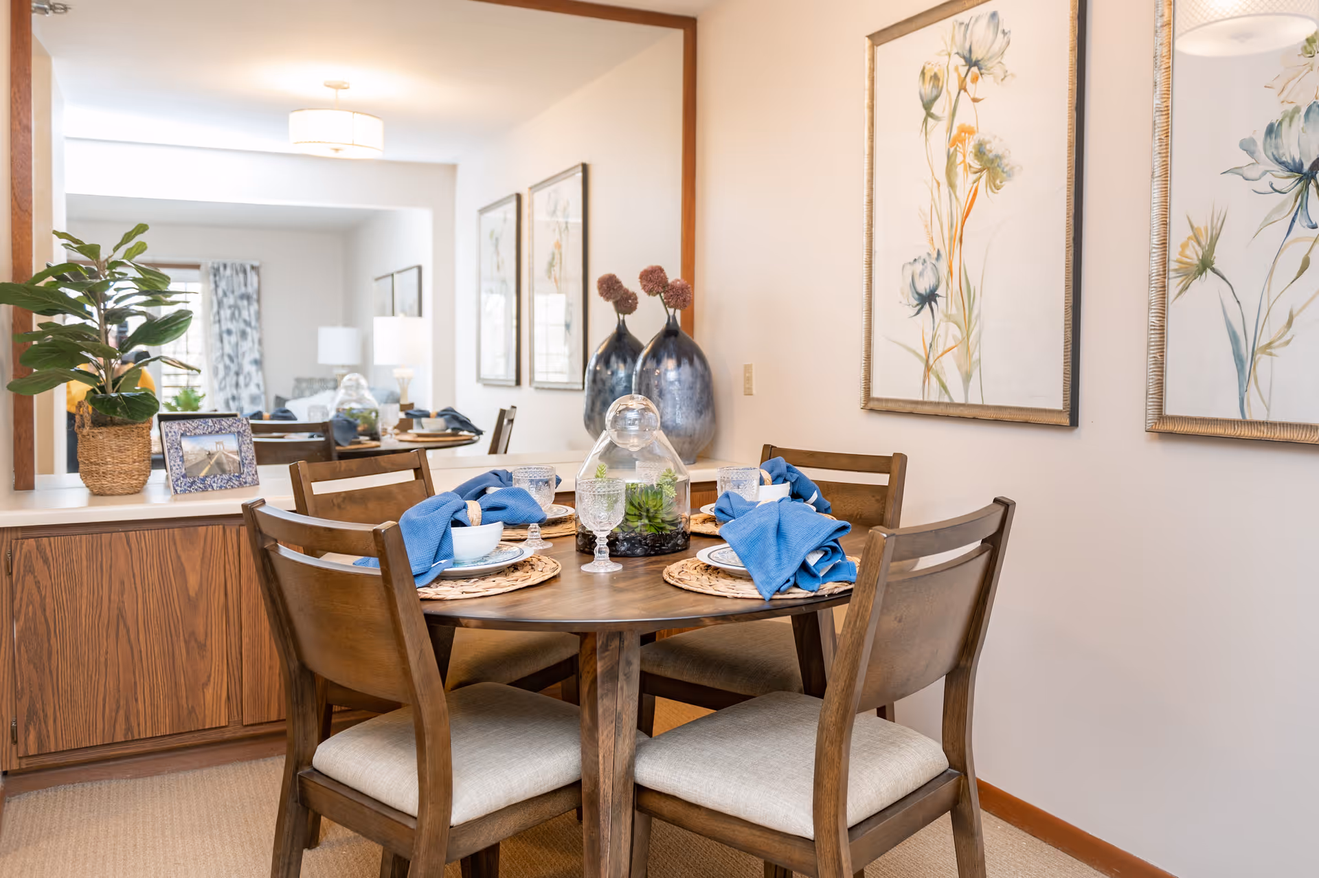 A cozy dining area with a round wooden table set for four, featuring blue napkins, glassware, and white dishes. The room has wooden chairs with beige cushions, a large mirror on the wall reflecting the adjacent living space, decorative vases, framed floral artwork, and a potted plant on a wooden sideboard.