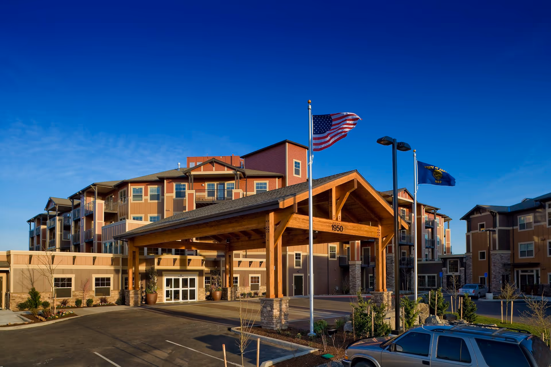 Exterior view of The Springs at Tanasbourne senior living facility showing a large covered entrance with wooden beams, two flagpoles with the American flag and the State of Oregon flag, a multi-story building with balconies, and a parked SUV in the foreground under a clear blue sky.