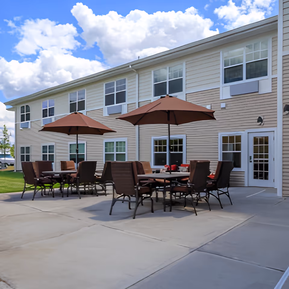 Outdoor patio with tables, chairs, and umbrellas in front of a two-story assisted living building.