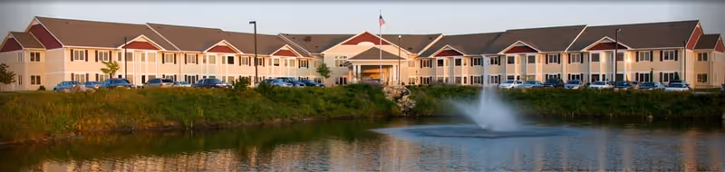 Wide exterior view of a large senior living facility building with beige and red accents, multiple windows, and a central entrance. In front of the building is a pond with a water fountain, surrounded by greenery and a parking lot with cars.