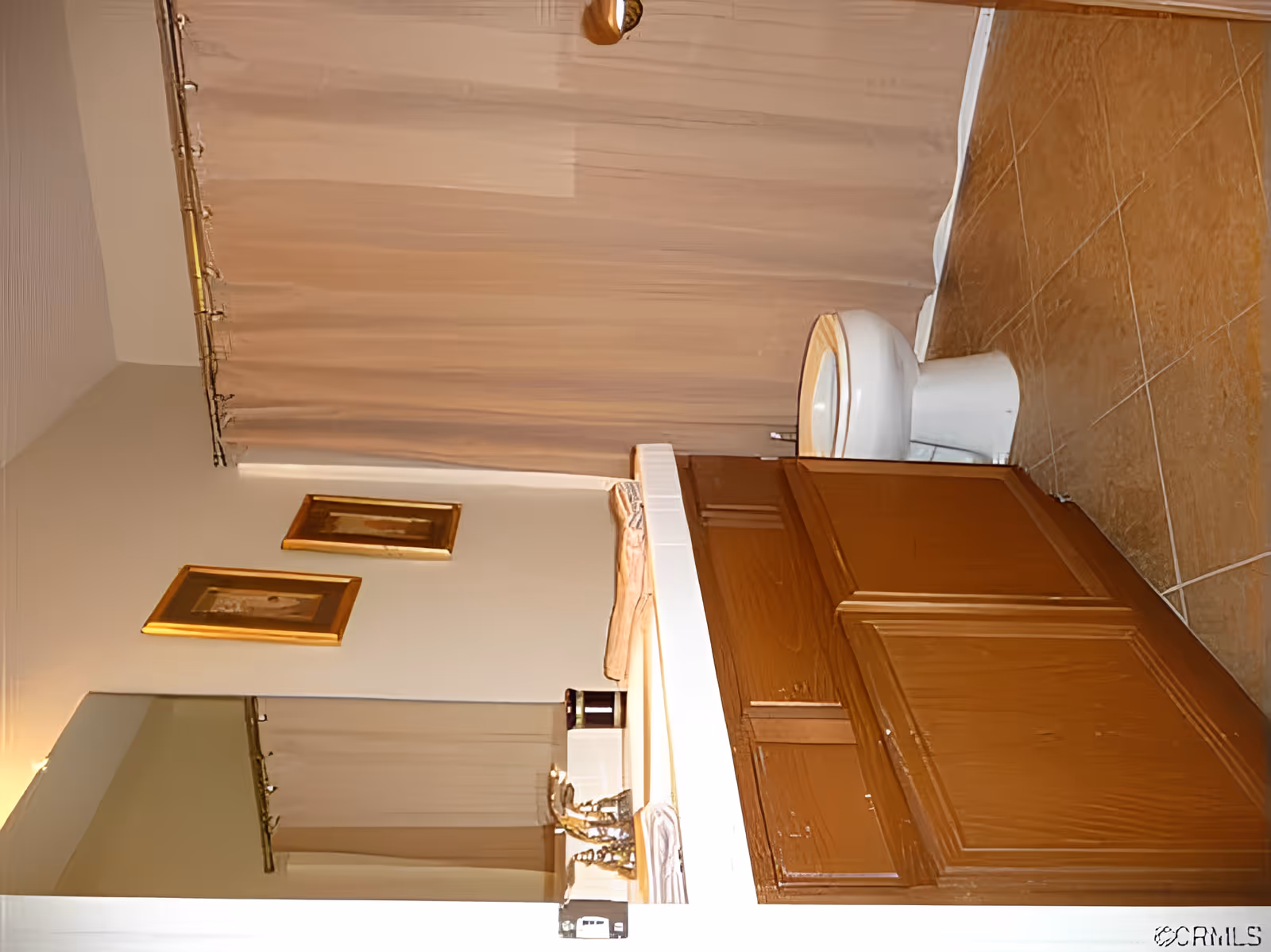 Bathroom with a wooden vanity cabinet, white sink, toilet, beige tiled floor, beige shower curtain, and two framed pictures on the wall above the sink.