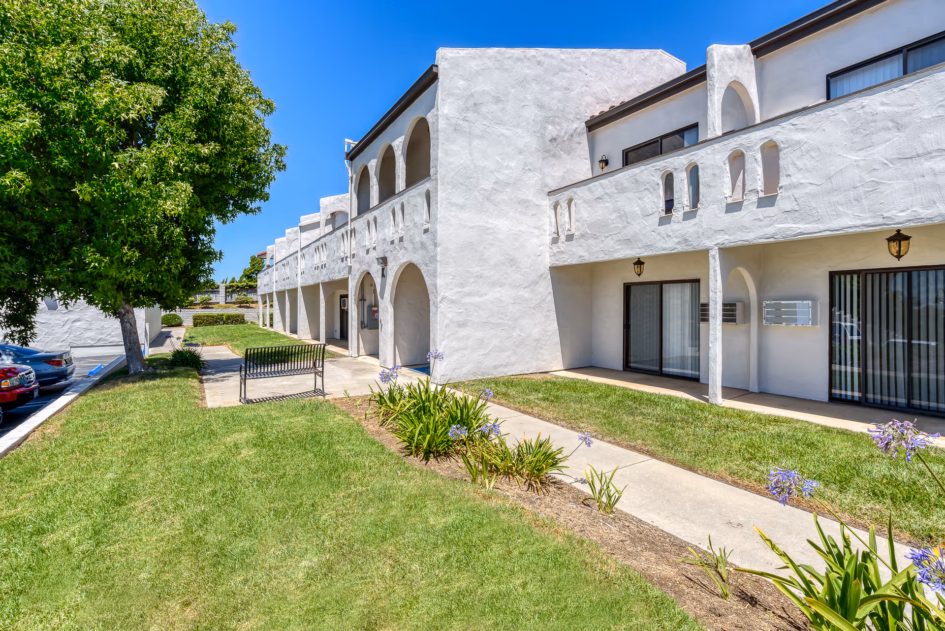 Exterior view of a white stucco building with arches and balconies at Rancho Vista Senior Living. There is a green lawn with a sidewalk, some flowering plants, a bench, and a tree. Several cars are parked along the left side under a clear blue sky.