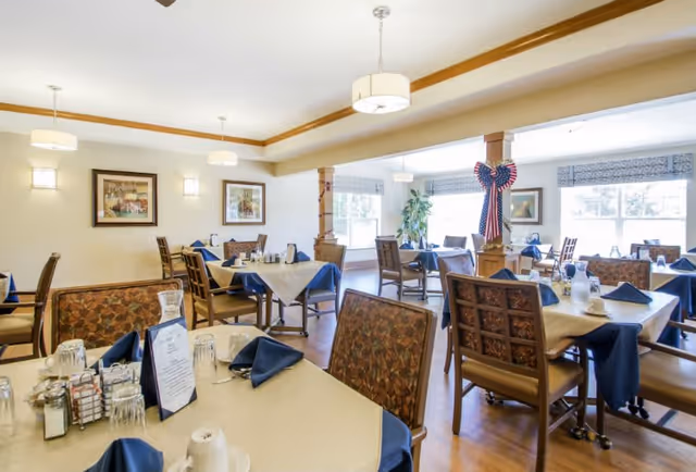 A bright and spacious dining room with multiple tables covered in beige tablecloths and blue napkins. Each table is set with glasses, cups, and condiments. The room features wooden chairs with patterned cushions, large windows letting in natural light, framed artwork on the walls, and decorative columns, one adorned with a patriotic bow.