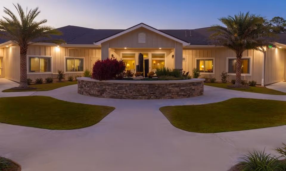 Exterior view of Cardinal Court Assisted Living & Memory Care building at dusk with well-lit windows, palm trees, and a circular stone planter in front surrounded by neatly trimmed grass and paved walkways.