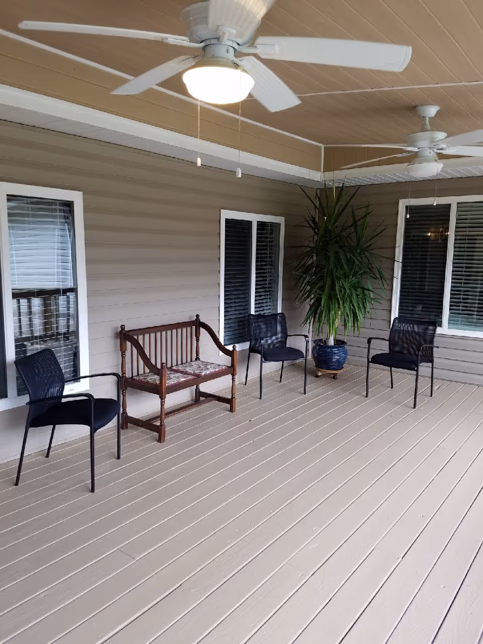 Covered outdoor patio area with beige wooden flooring and walls, featuring two ceiling fans with lights, three black mesh chairs, a wooden bench with a floral cushion, and a large potted plant near two windows with white blinds.