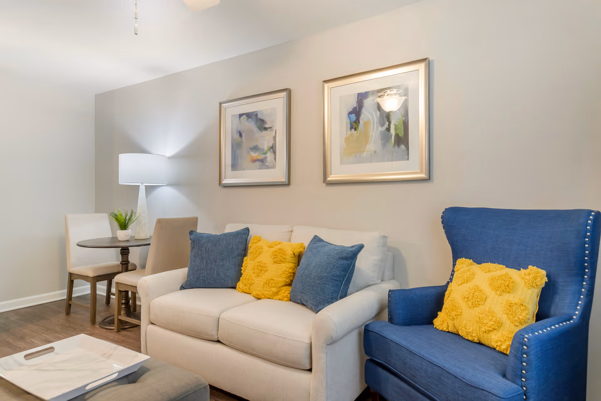 A cozy living room area with a beige loveseat adorned with two blue pillows and one yellow textured pillow. Next to it is a blue armchair with a matching yellow textured pillow. Behind the loveseat are two abstract paintings in silver frames hung on a light gray wall. To the left, there is a small round wooden table with two beige chairs and a white table lamp with a white shade. A small potted plant is on the table. The floor is wooden, and a white tray is visible on a gray ottoman in the foreground.