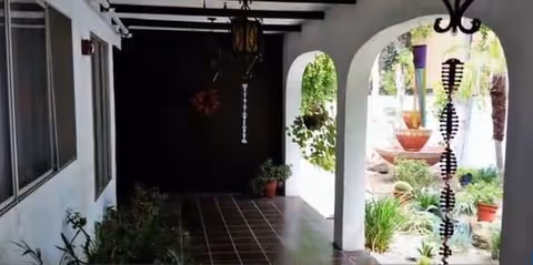 Covered outdoor walkway with white walls and dark tiled floor, featuring arched openings on the right side that look out onto a garden area with plants and a fountain. There are hanging decorative elements and potted plants along the walkway.