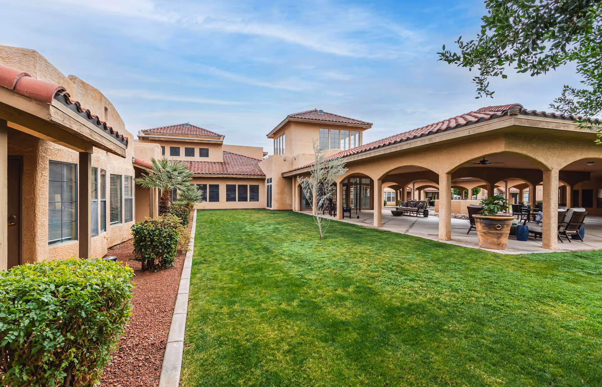 Outdoor courtyard area of a senior living facility with green grass, shrubs, and trees. The buildings have beige stucco walls and red tile roofs. There is a covered patio with arches and outdoor seating including chairs and tables.