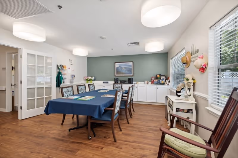 A well-lit room with a long table covered by a blue tablecloth surrounded by wooden chairs with floral cushions. The room has wooden flooring, white walls, and large windows with blinds. There is a white cabinet against a green accent wall with a framed picture above it. On the right side, there is a small white table with decorative hats and a rocking chair with a green cushion. The room appears clean and organized, suitable for meetings or activities.