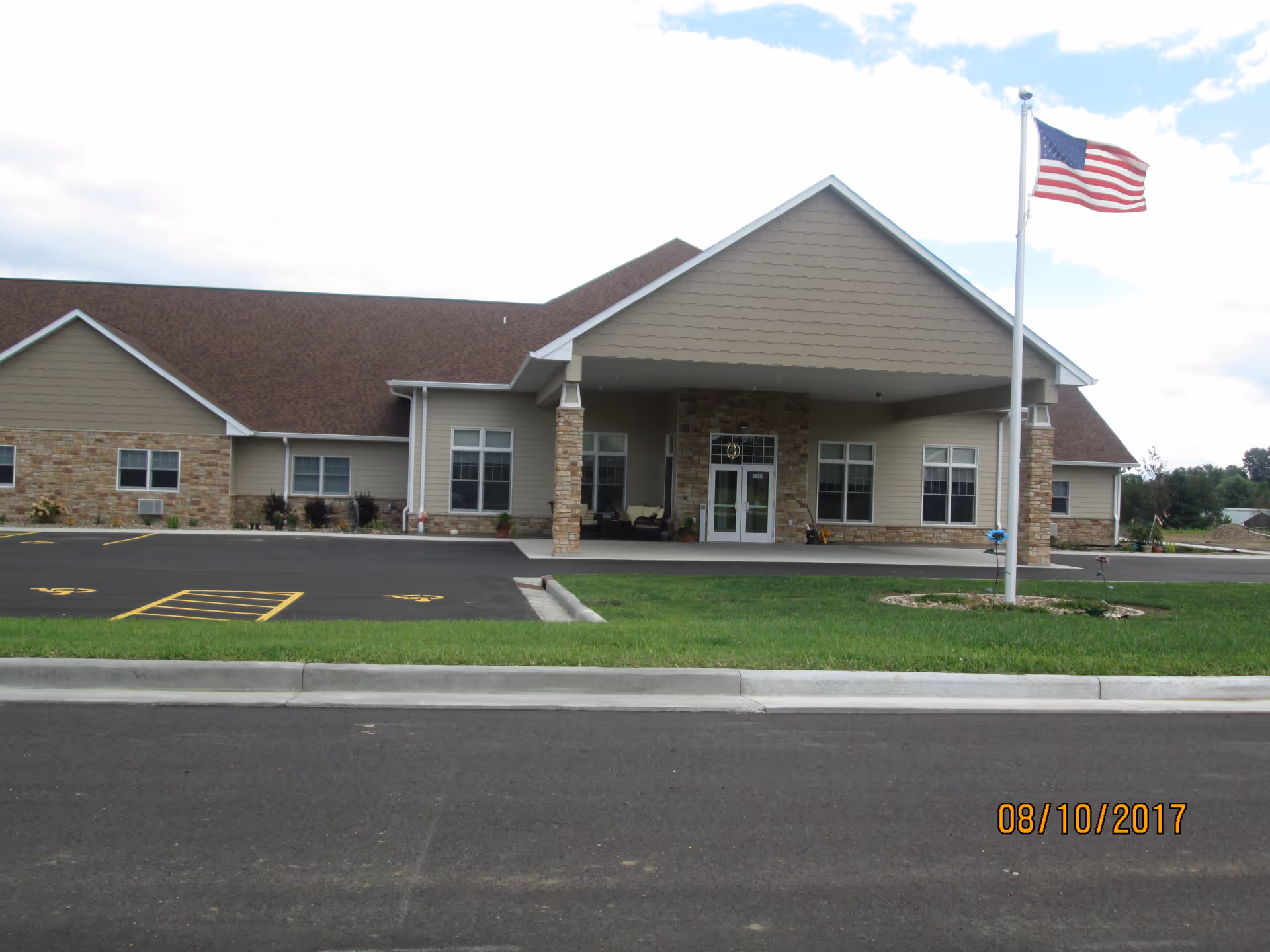 Front exterior view of a single-story building with a covered entrance supported by stone pillars. The building has beige siding and stone accents, a brown shingled roof, and several windows. An American flag on a flagpole is positioned on the right side of the entrance. The parking lot and a grassy area are visible in front of the building under a partly cloudy sky.