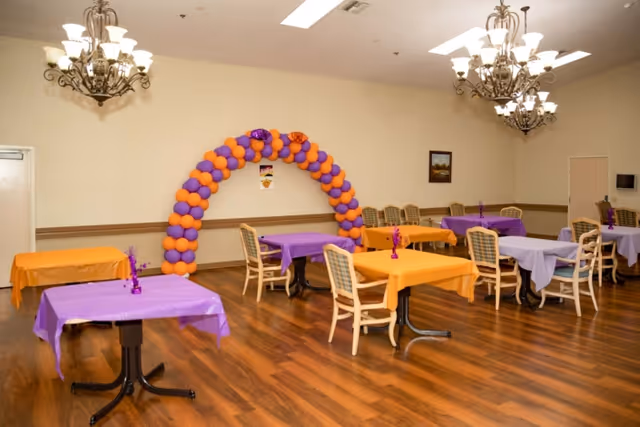 A decorated dining/activity room with tables draped in purple and orange cloths, chairs, and a purple-and-orange balloon arch along the back wall.