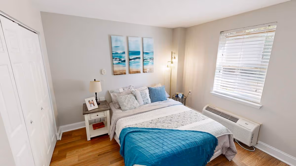 A cozy bedroom with a neatly made bed featuring white and blue bedding. There is a nightstand with a lamp and a framed photo on the left side of the bed. Above the bed, there are three pieces of wall art depicting a beach scene. A window with white blinds is on the right wall, and a heating/cooling unit is below the window. The room has wooden flooring and light-colored walls.