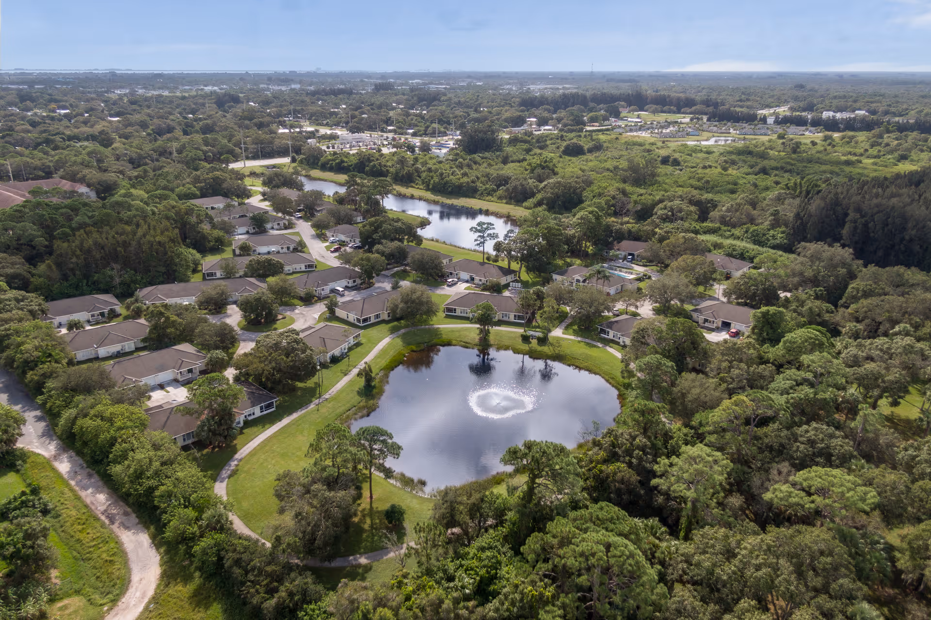 Aerial view of Lake Forest Park senior living community showing multiple single-story residential buildings surrounding a central pond with a water fountain, surrounded by lush greenery and trees under a partly cloudy sky.
