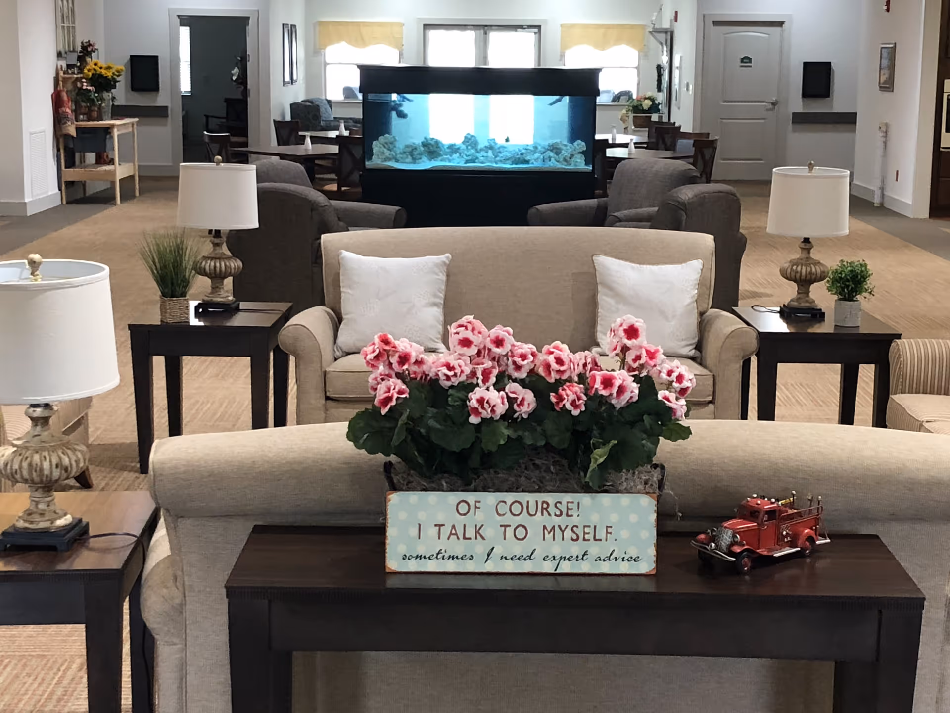 A cozy living room area in a senior living facility featuring beige sofas with white pillows, dark wooden side tables with lamps, a decorative plant with pink flowers, and a small red toy fire truck on a table. In the background, there is a large aquarium and additional seating with armchairs and tables. The room has a warm and inviting atmosphere with neutral tones and soft lighting.