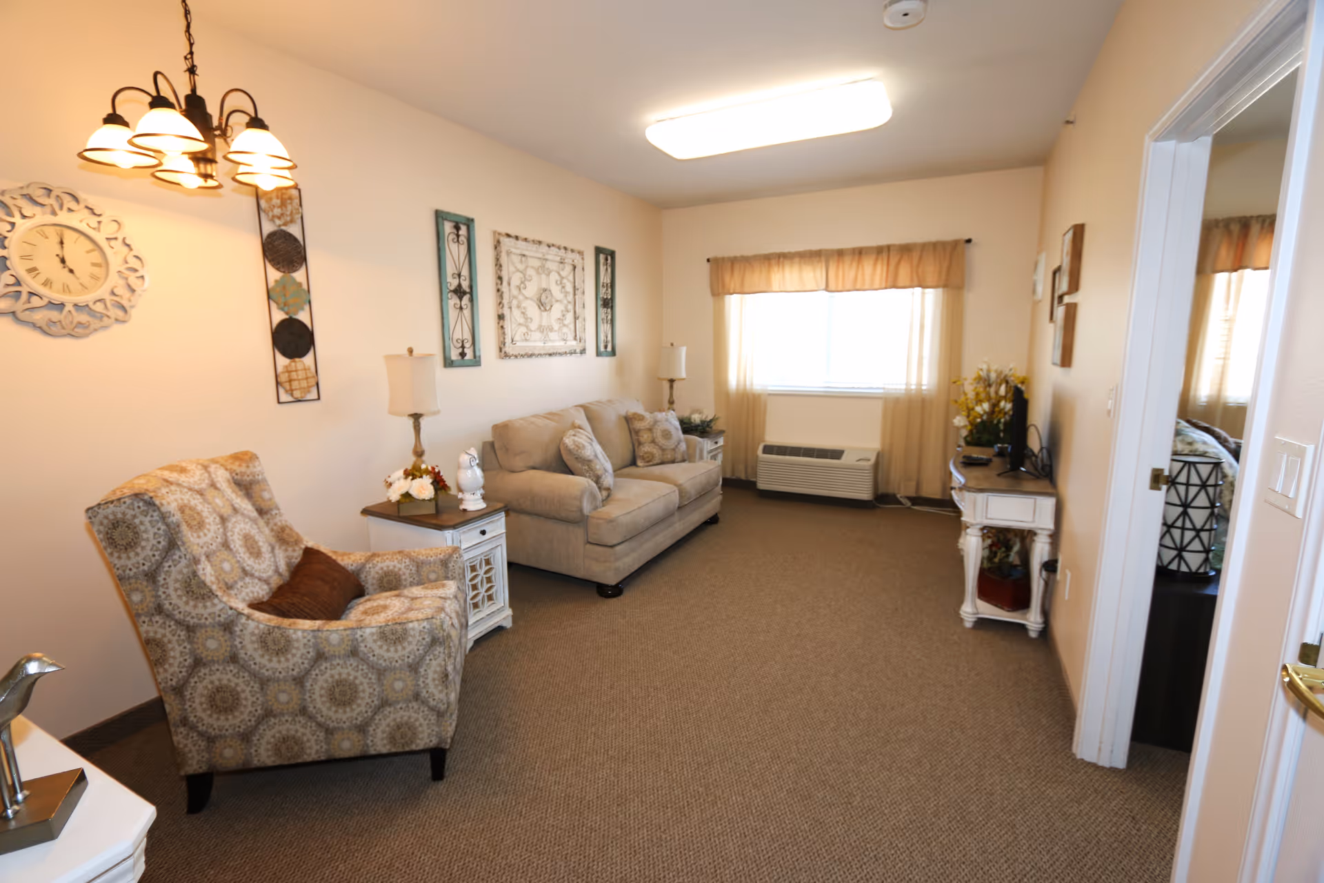 Bright living room with a patterned armchair, beige sofa, side tables, wall art, and a window air conditioner.