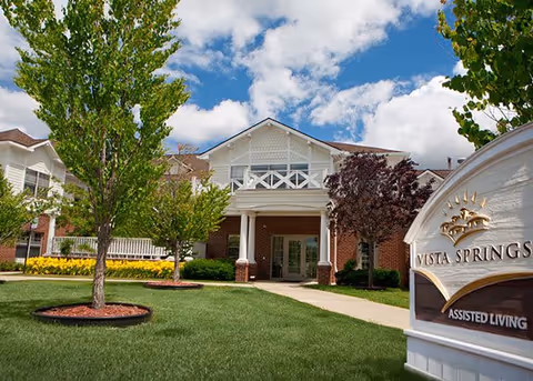 Exterior view of a senior living facility with a well-maintained lawn, trees, and a clear blue sky with some clouds. The building has a brick facade with white trim and a covered entrance supported by columns. A sign in the foreground reads 'Vista Springs Assisted Living.'