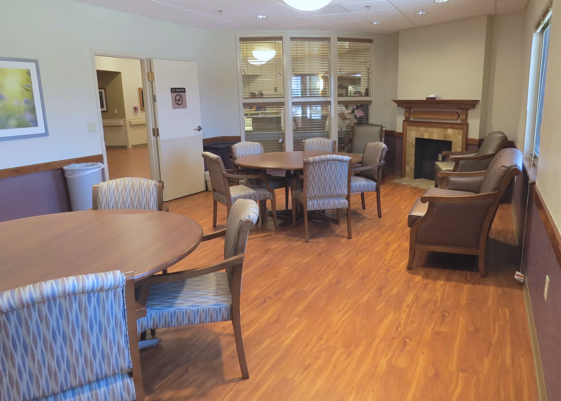 A cozy common area in a nursing and rehabilitation center featuring round wooden tables with patterned cushioned chairs, a fireplace with a wooden mantle, and several armchairs along the wall. The room has wood flooring, beige walls, and a window with blinds. A door is open to a hallway, and a framed picture hangs on the wall.