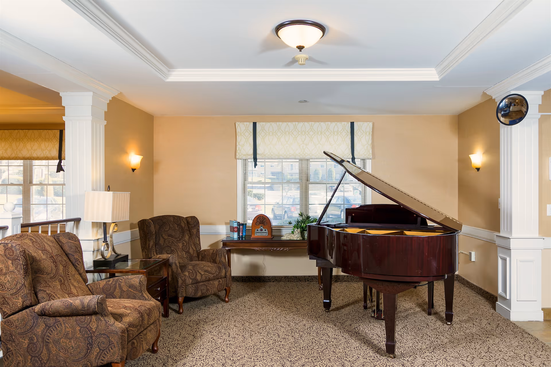 A cozy interior room with two patterned armchairs, a side table with a lamp, a grand piano with its lid open, and a window with blinds and a decorative valance. The walls are beige with white trim, and there is a ceiling light fixture and wall sconces providing warm lighting.