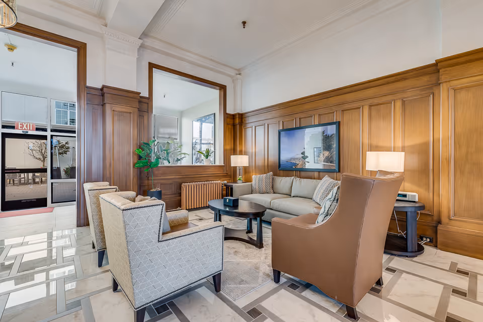 A bright and elegant senior living facility lounge area with wood-paneled walls, a large window, and a glass door leading outside. The room features a beige sofa with patterned cushions, two upholstered armchairs, a brown leather armchair, a round coffee table, side tables with lamps, and a potted plant. The floor has a polished tile design, and a large framed picture hangs on the wall above the sofa.