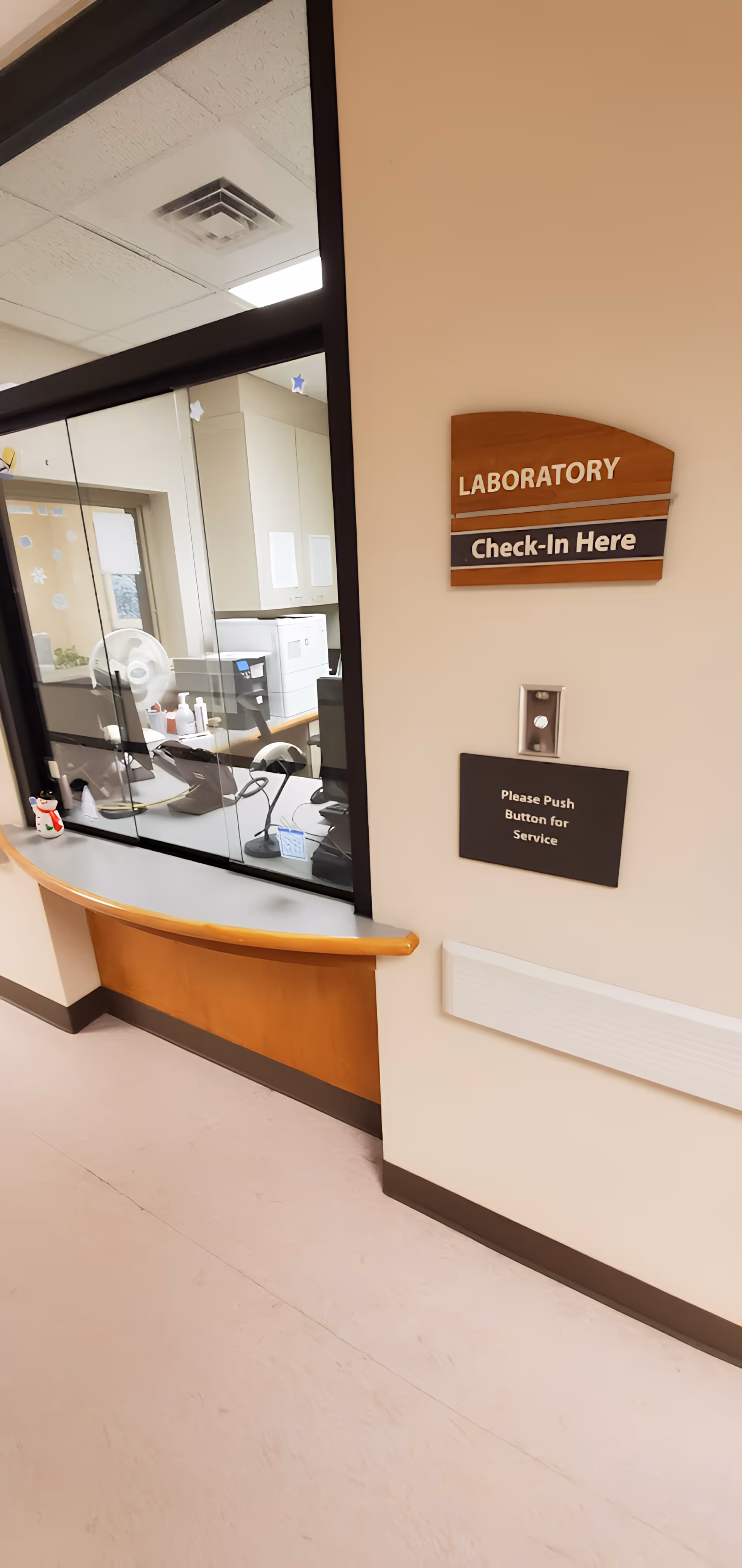 Interior view of a laboratory check-in window at Cox Medical Centers Meyer Orthopedic And Rehab. The window has a glass partition with a counter below it. Behind the glass, there are office supplies, a computer, a fan, and cabinets. On the wall next to the window, there is a wooden sign that reads 'LABORATORY Check-In Here' and a smaller sign below it that says 'Please Push Button for Service'.