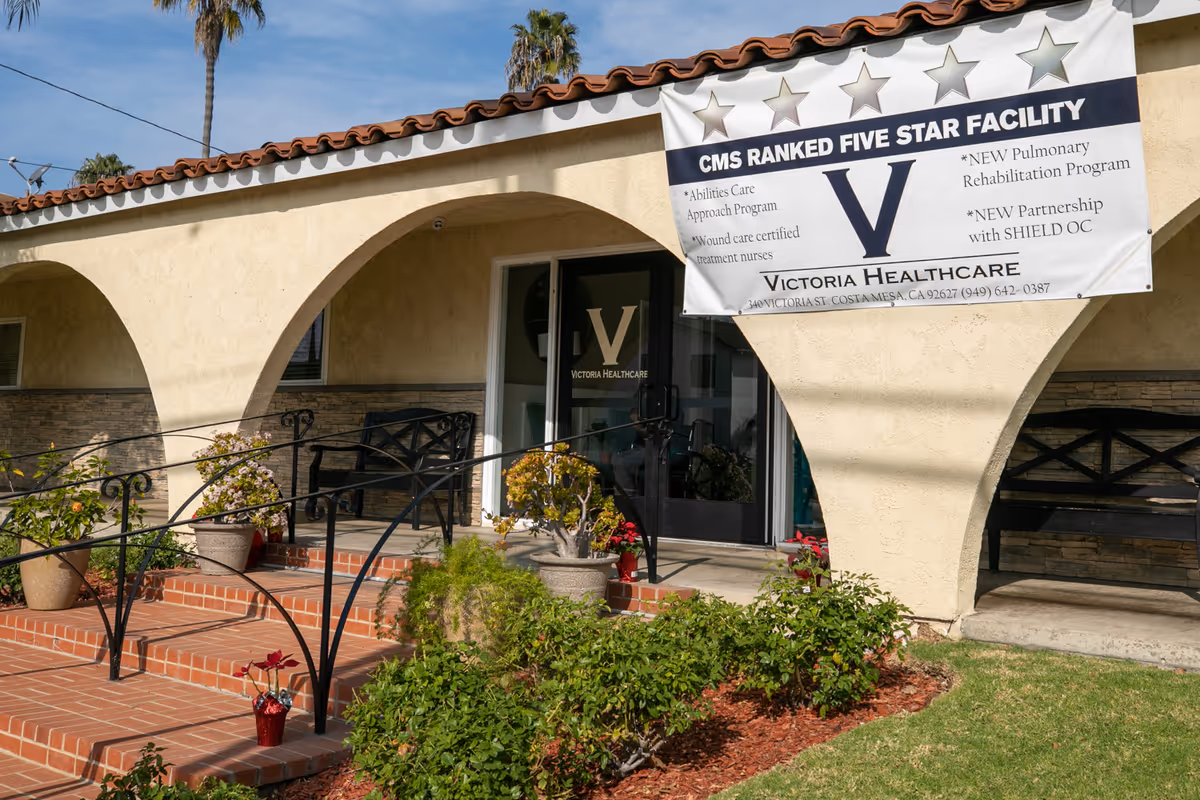 Entrance of Victoria Healthcare and Rehabilitation facility with a banner displaying CMS ranked five star facility and various program highlights. The building has a beige stucco exterior with arched doorways, potted plants, benches, and a brick walkway with a black metal railing.
