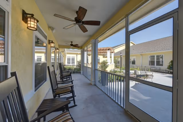 A screened-in porch area with several wooden rocking chairs lined up along a yellow exterior wall. The porch has ceiling fans and wall-mounted lantern-style lights. Outside the screened area, there is a courtyard with benches, greenery, and a pergola structure under a clear blue sky.