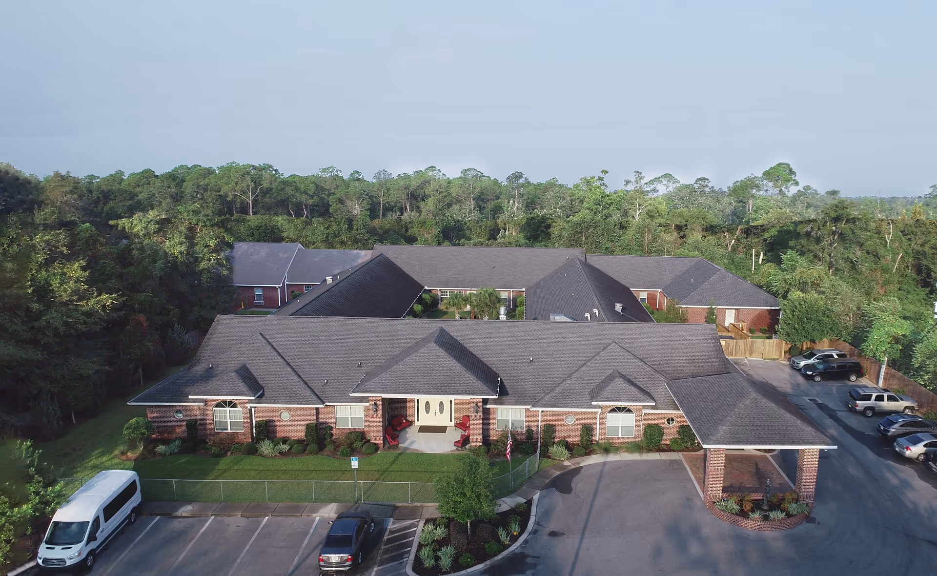 Aerial view of a single-story brick senior living building with a covered entrance, parking lot, and surrounding trees.