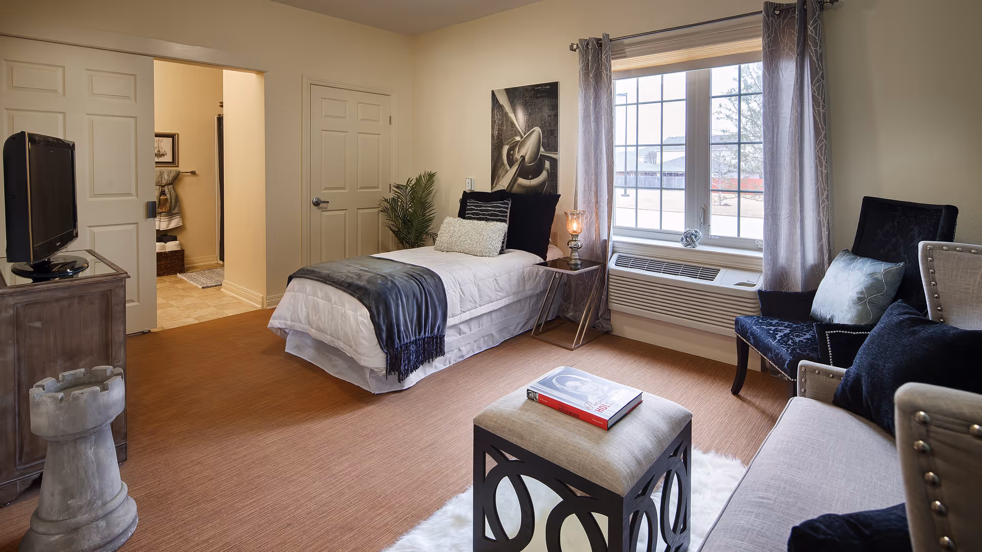 A cozy bedroom with a single bed dressed in white linens and a dark throw blanket. Next to the bed is a small side table with a lamp and a decorative plant. There is a large window with curtains letting in natural light. The room also features a cushioned ottoman with a book on top, a dark upholstered chair, and a light-colored sofa with pillows. A TV sits on a wooden cabinet near the door, and an open doorway leads to a bathroom.