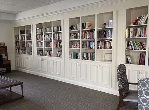 A room with a long wall of built-in white bookshelves filled with books and some decorative items. There is a gray carpeted floor and two patterned chairs partially visible on the right side. The room has a calm and organized atmosphere.