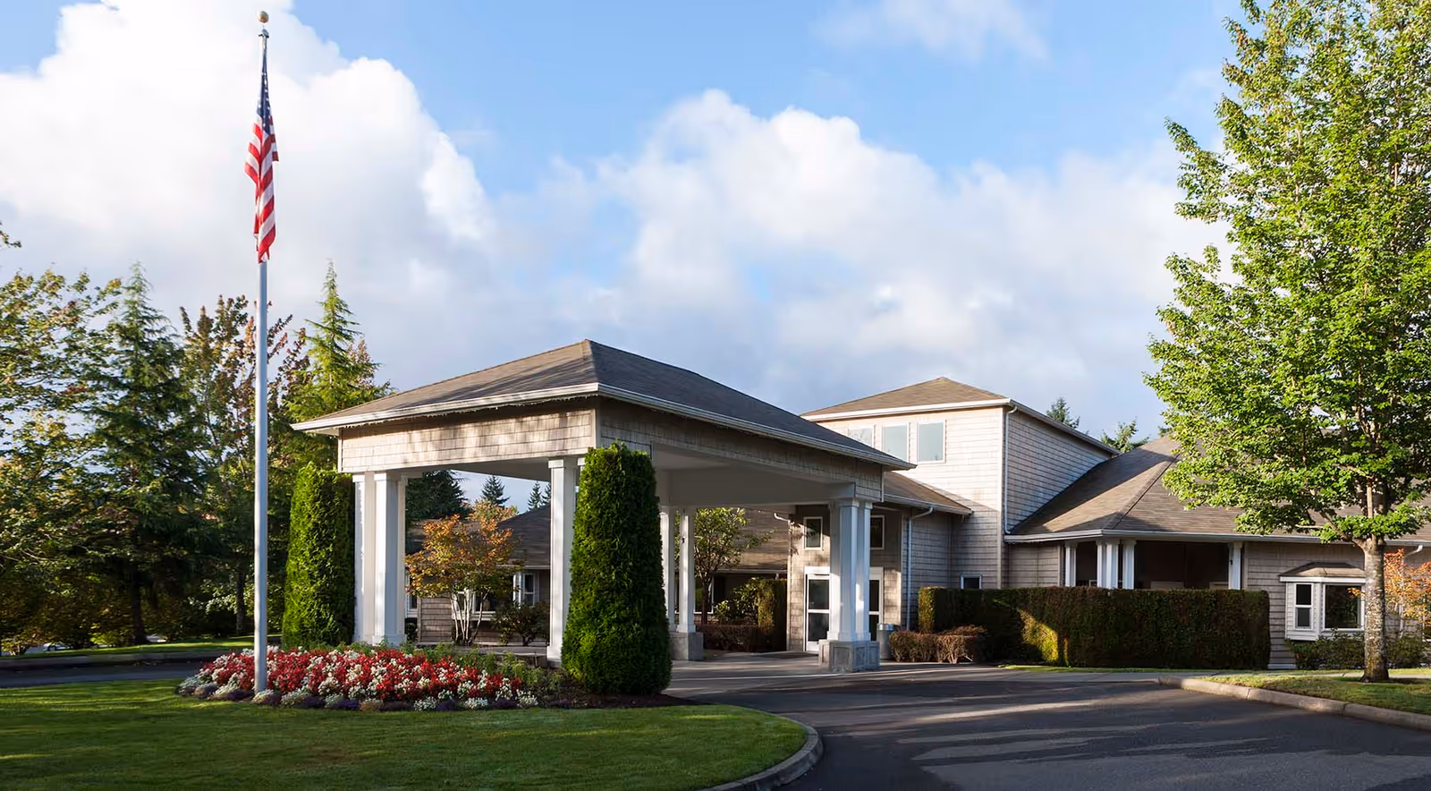 Exterior view of Avalon Federal Way Care Center showing the entrance with a covered driveway, an American flag on a flagpole, well-maintained landscaping with flowers and trees, and a partly cloudy sky.