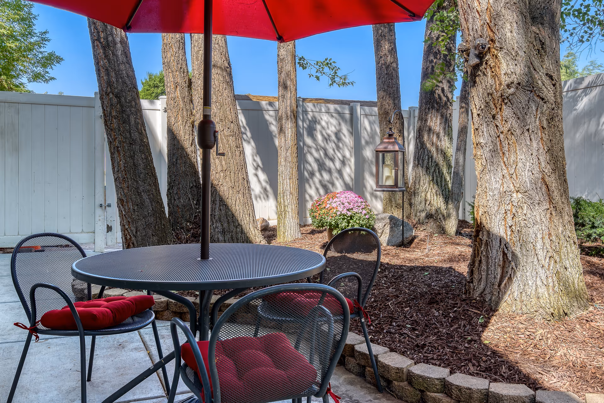 Outdoor patio area with a round metal table and four matching chairs, each with red cushions. A large red umbrella provides shade. The patio is surrounded by tall trees, a white fence, a flower pot with pink flowers, and a hanging lantern with candles.