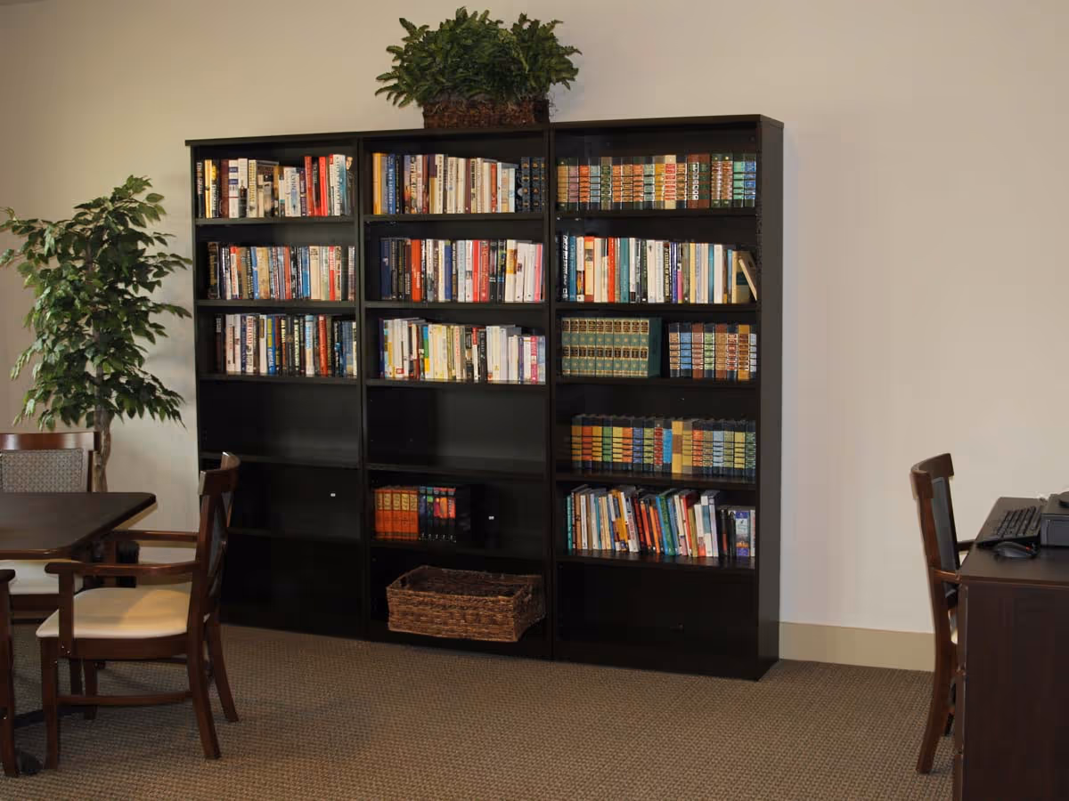 A quiet reading or study area with a large black bookshelf filled with books, a green potted plant on top of the bookshelf, a small green leafy plant to the left, a wooden table with chairs, and a desk with a computer on the right side.