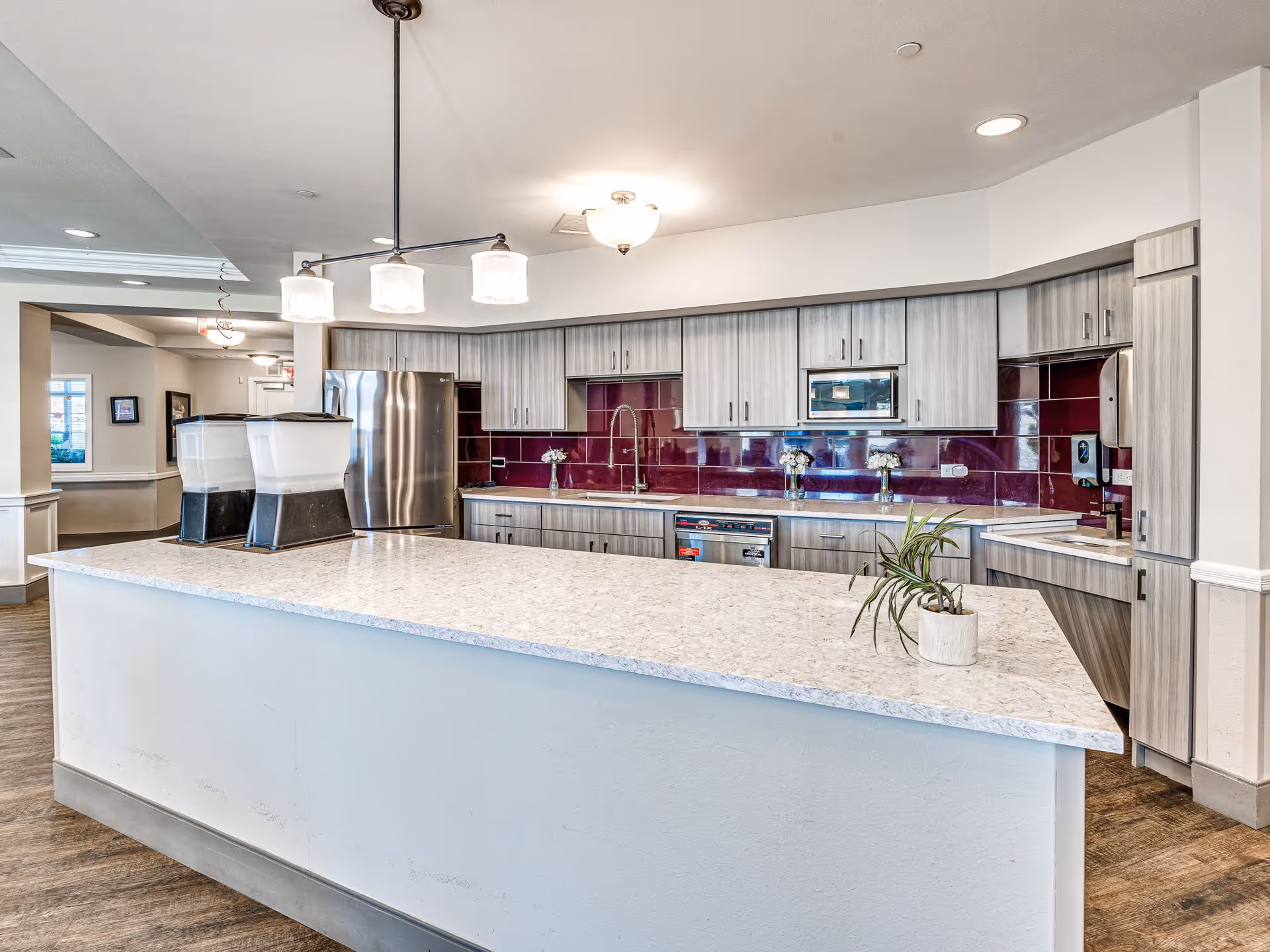 A modern kitchen area in a senior living facility featuring a large white marble countertop island with two beverage dispensers and a small potted plant. The back wall has light wood cabinets, a stainless steel refrigerator, a microwave, and a dishwasher. The backsplash is a glossy dark red tile. The floor is wood, and the ceiling has recessed lighting and a hanging light fixture above the island.