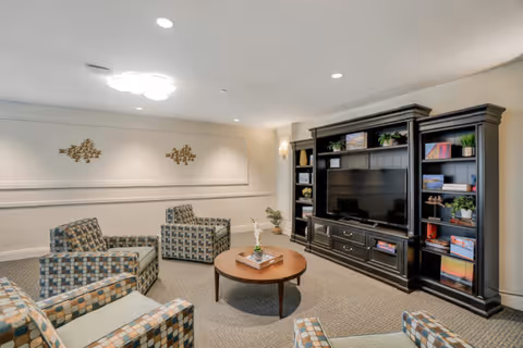 A cozy common living room with patterned armchairs arranged around a round coffee table facing a large dark wood entertainment center with a television and shelves.