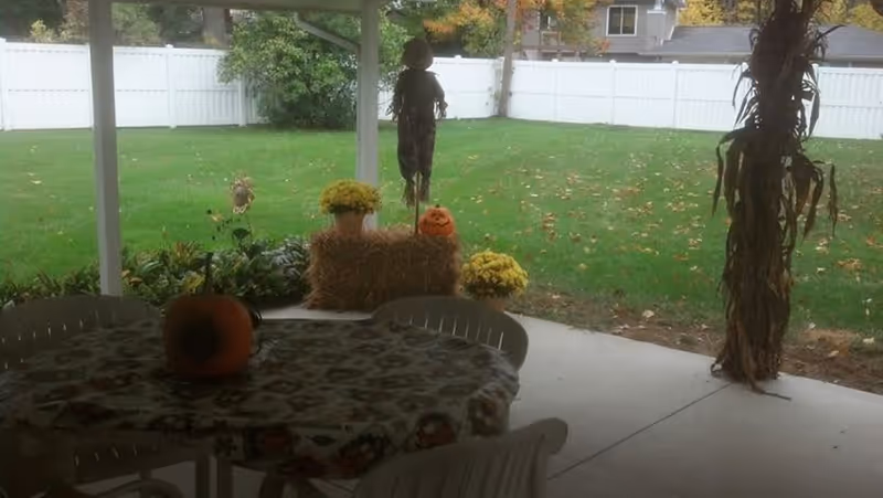 Covered outdoor patio area with a table covered by a patterned tablecloth and a pumpkin centerpiece. The patio overlooks a green lawn enclosed by a white fence. There are autumn decorations including a scarecrow, hay bales, yellow flowers, and a small pumpkin with a carved face.