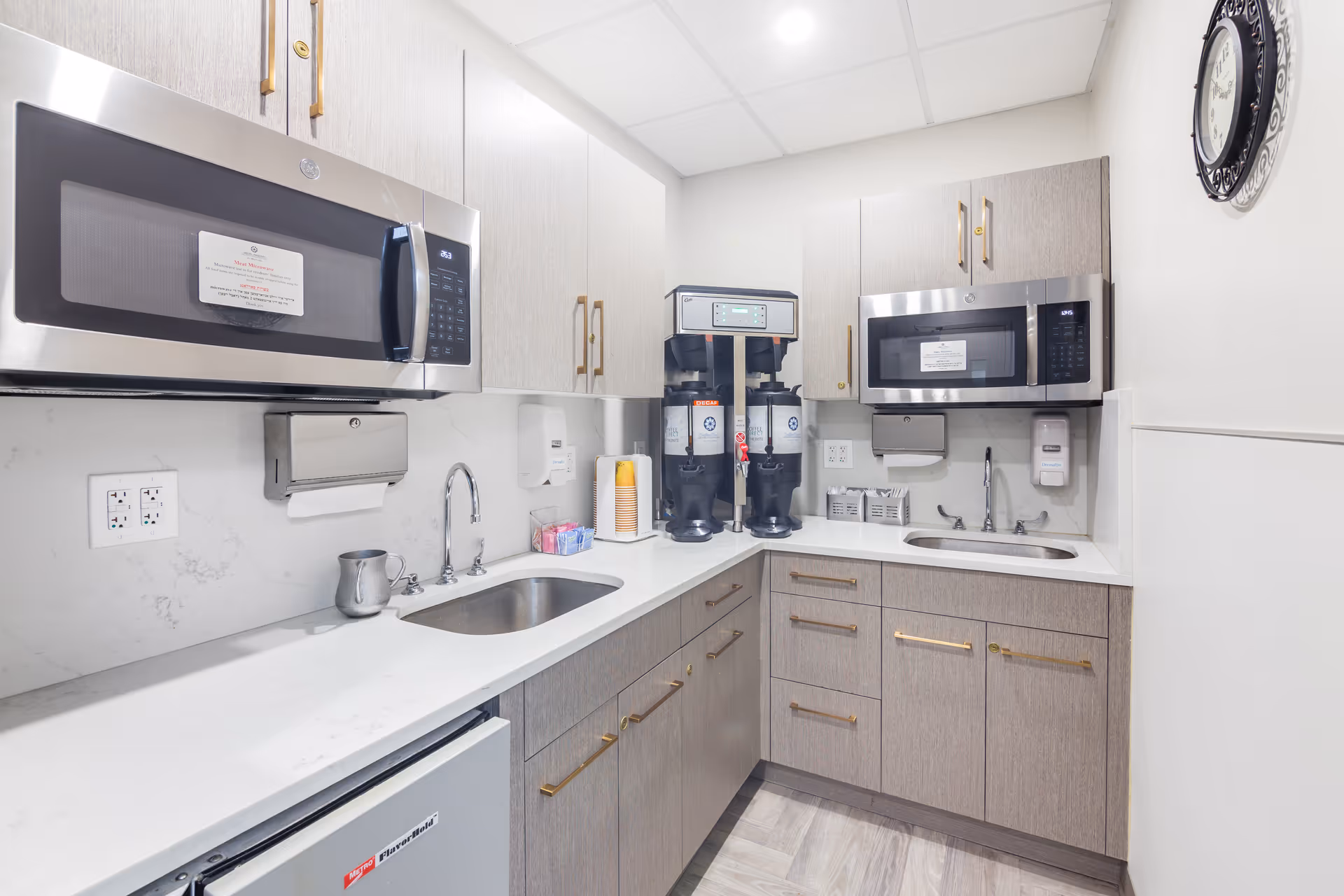 A small kitchenette area with two stainless steel microwaves mounted above white countertops. The countertops have two sinks, a coffee machine with two large coffee dispensers, a paper towel dispenser, a soap dispenser, and various small items like cups and sugar packets. The cabinets are light wood with gold handles, and there is a clock on the wall to the right.