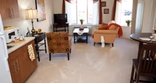 A bright living area in a retirement facility featuring a small kitchenette with microwave and cabinets on the left, a patterned armchair, a TV on a stand near a window with curtains, a beige sofa with an orange throw, a small coffee table with a decorative item, and a round dining table with chairs on the right.