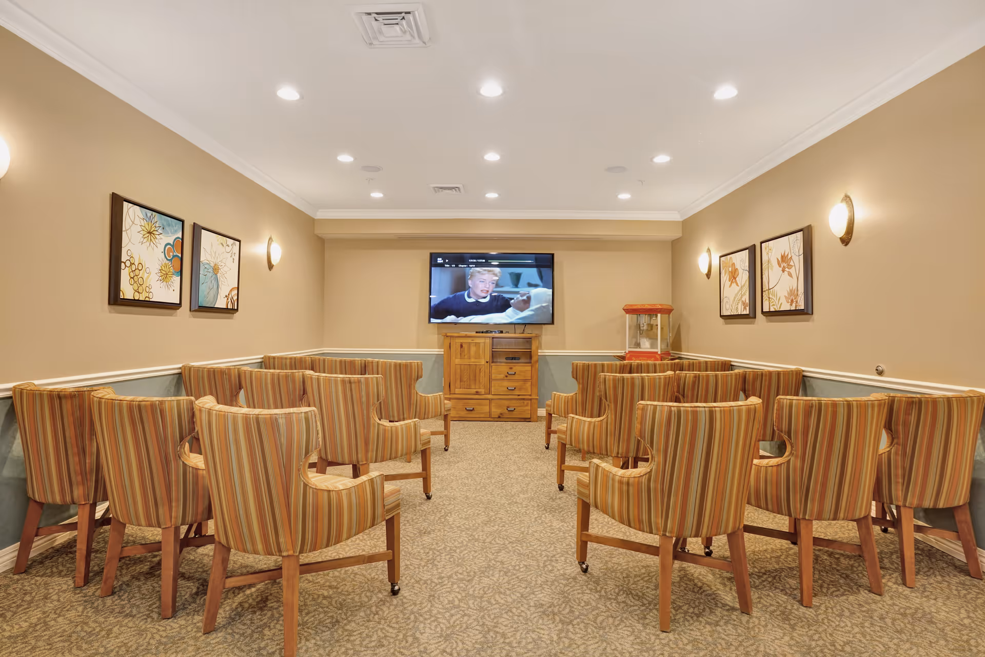 A small theater room with two rows of striped upholstered chairs facing a wall-mounted flat screen TV. The walls are beige with framed floral artwork and wall sconces providing light. A wooden cabinet and a popcorn machine are positioned below the TV.