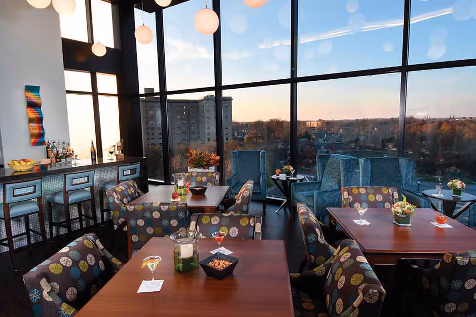 A modern lounge area with large floor-to-ceiling windows offering a view of a cityscape at sunset. The room features several tables with patterned chairs, a bar with stools, and comfortable armchairs near the windows. Drinks and small bowls of snacks are placed on the tables.