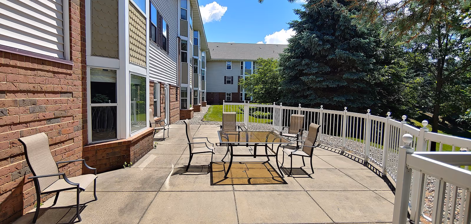 Outdoor patio area at Oak Meadows Senior Living with a glass-top table and six chairs on a concrete surface, surrounded by a white fence. The patio is adjacent to a building with brick and siding exterior, and there are trees and green grass in the background under a blue sky with some clouds.