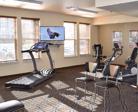 Bright exercise room with a treadmill, stationary bikes, chairs and a wall-mounted TV playing a fitness video.