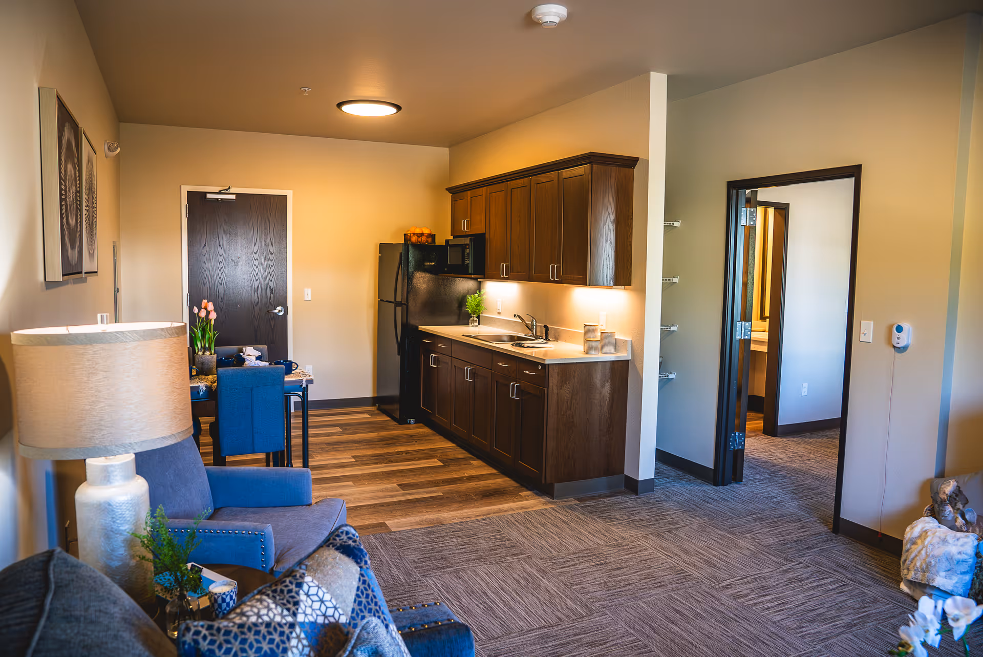 Interior view of a senior living facility apartment showing a small kitchen area with dark wooden cabinets, a black refrigerator, microwave, and sink. Adjacent to the kitchen is a dining table with blue chairs. The living area has a blue armchair, a lamp, and decorative pillows. There is a doorway leading to another room, and the flooring transitions from wood in the kitchen to carpet in the living area.