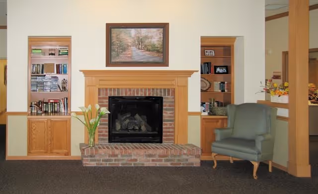 A cozy interior living area featuring a brick fireplace with a wooden mantel, flanked by built-in wooden shelves filled with books and decorative items. A framed painting of a forest path hangs above the fireplace. To the right of the fireplace is a green upholstered armchair, and a vase with white flowers is placed on the hearth.