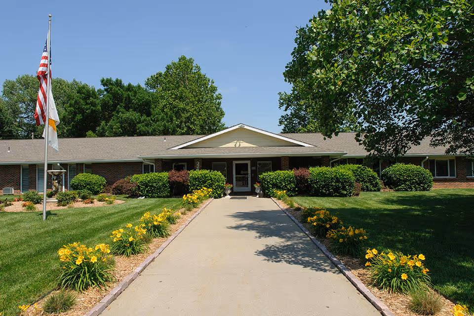 Walkway lined with yellow flowers leading to the entrance of a single-story brick senior living building with flags and landscaped lawns.