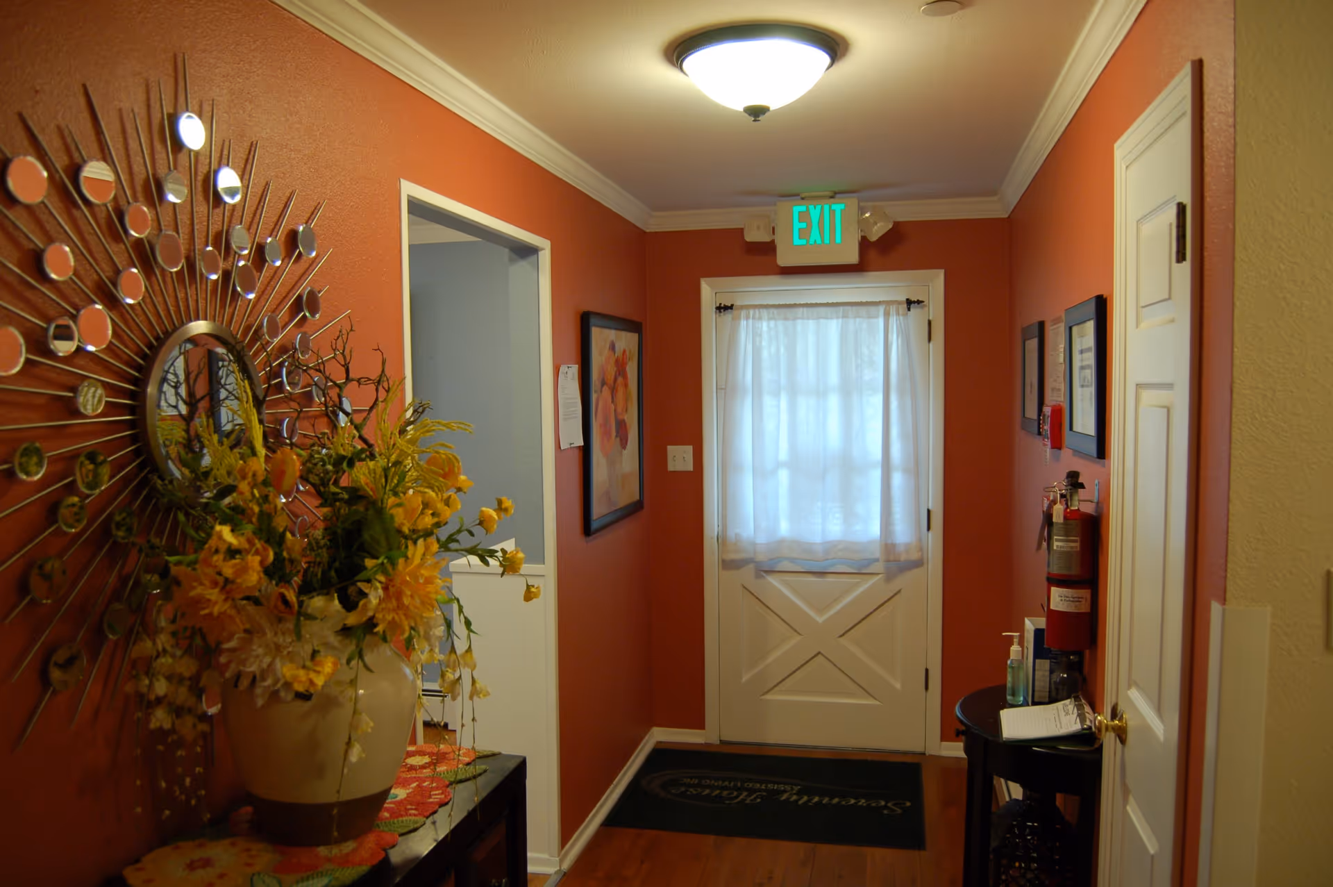 Interior hallway with orange walls, a decorative round mirror with sunburst design on the left wall, a large vase with yellow flowers on a table below the mirror, a white door with a sheer curtain and an illuminated green exit sign above it at the end of the hallway, framed artwork on the walls, a fire extinguisher mounted on the right wall, and a small table with hand sanitizer and papers.