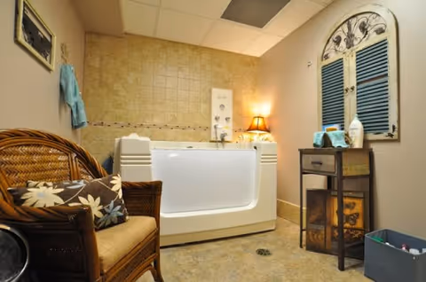 A cozy bathroom featuring a walk-in bathtub with a door, beige tiled walls, a wicker chair with floral cushions, a small wooden side table with towels and toiletries, a decorative wall piece resembling a window shutter, and a lit table lamp providing warm lighting.