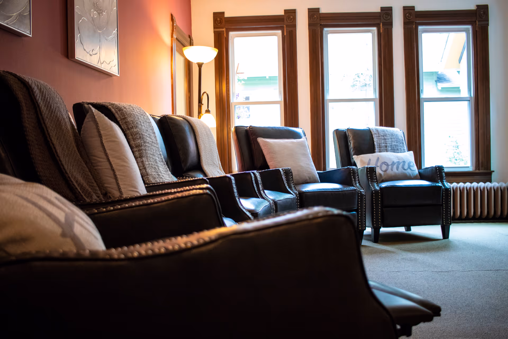 A cozy living room with several dark leather armchairs lined up, each with cushions and throws. The room has three tall windows with wooden frames letting in natural light, a floor lamp with two lights, and a radiator beneath the windows.