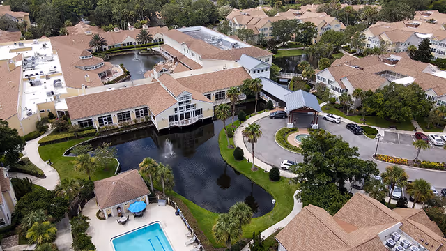 Aerial view of Vicar's Landing at Sawgrass senior living facility showing multiple buildings with tan roofs surrounding a pond with a fountain, a swimming pool with blue umbrellas, palm trees, and a circular driveway with parked cars.